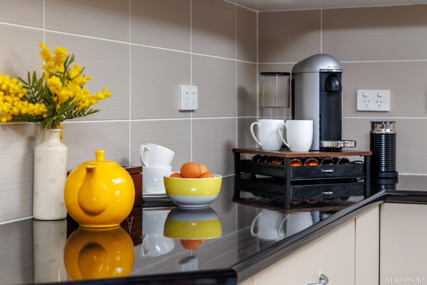 Et moderne køkken med en sort bordplade, kaffemaskine, elkedel, en skål med appelsiner og en vase med gule blomster.