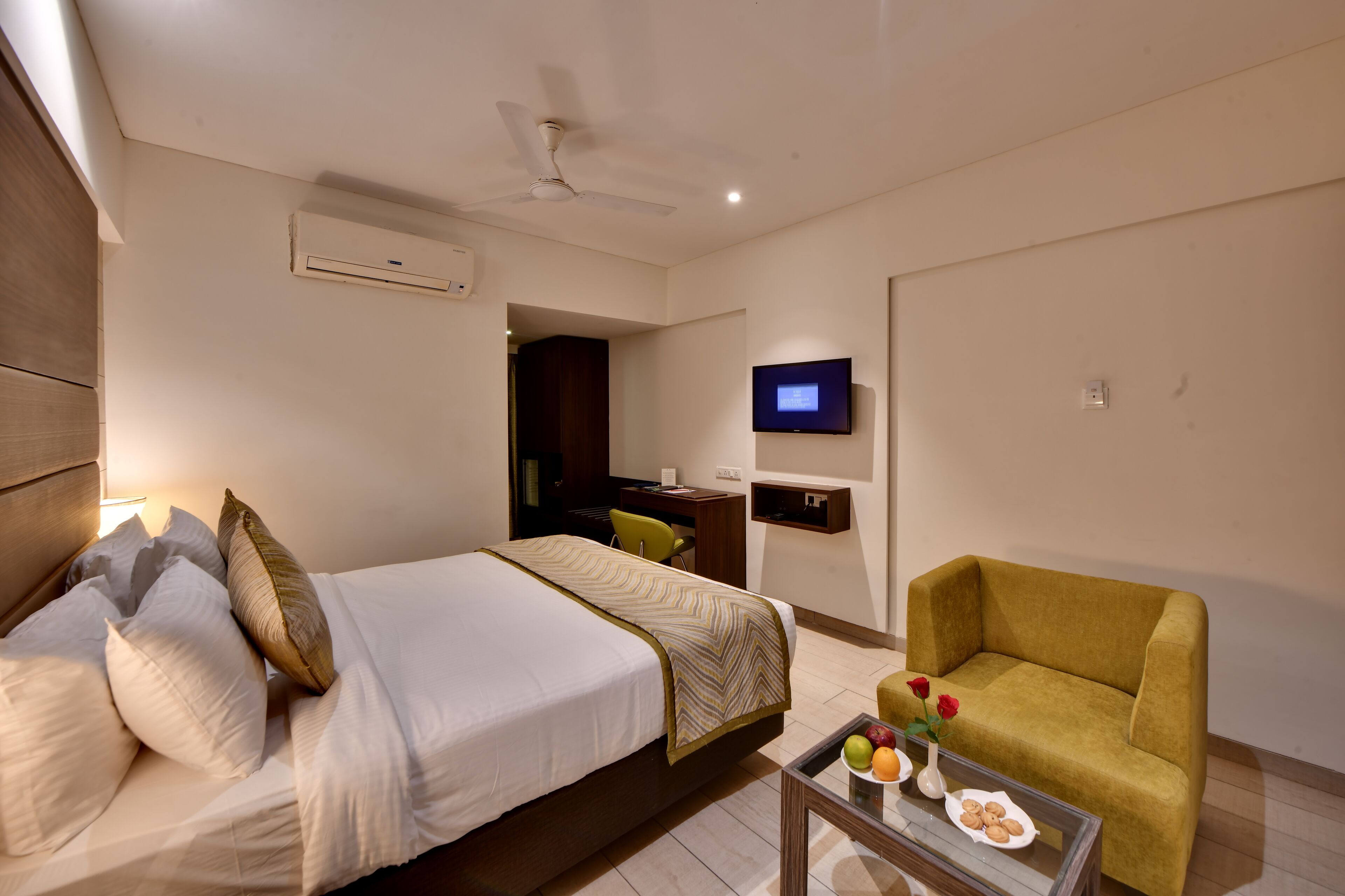 A hotel room with a bed, a sofa, a TV, and a small table with fruit and cookies.