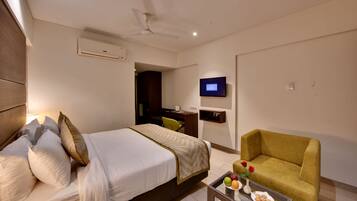 A hotel room with a bed, a sofa, a TV, and a small table with fruit and cookies.