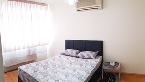 Une chambre à coucher comprenant un lit, une table de chevet, un luminaire suspendu et une fenêtre avec des rideaux légers.