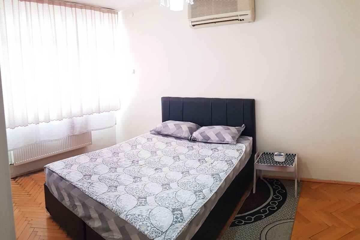 Une chambre à coucher comprenant un lit, une table de chevet, un luminaire suspendu et une fenêtre avec des rideaux légers.