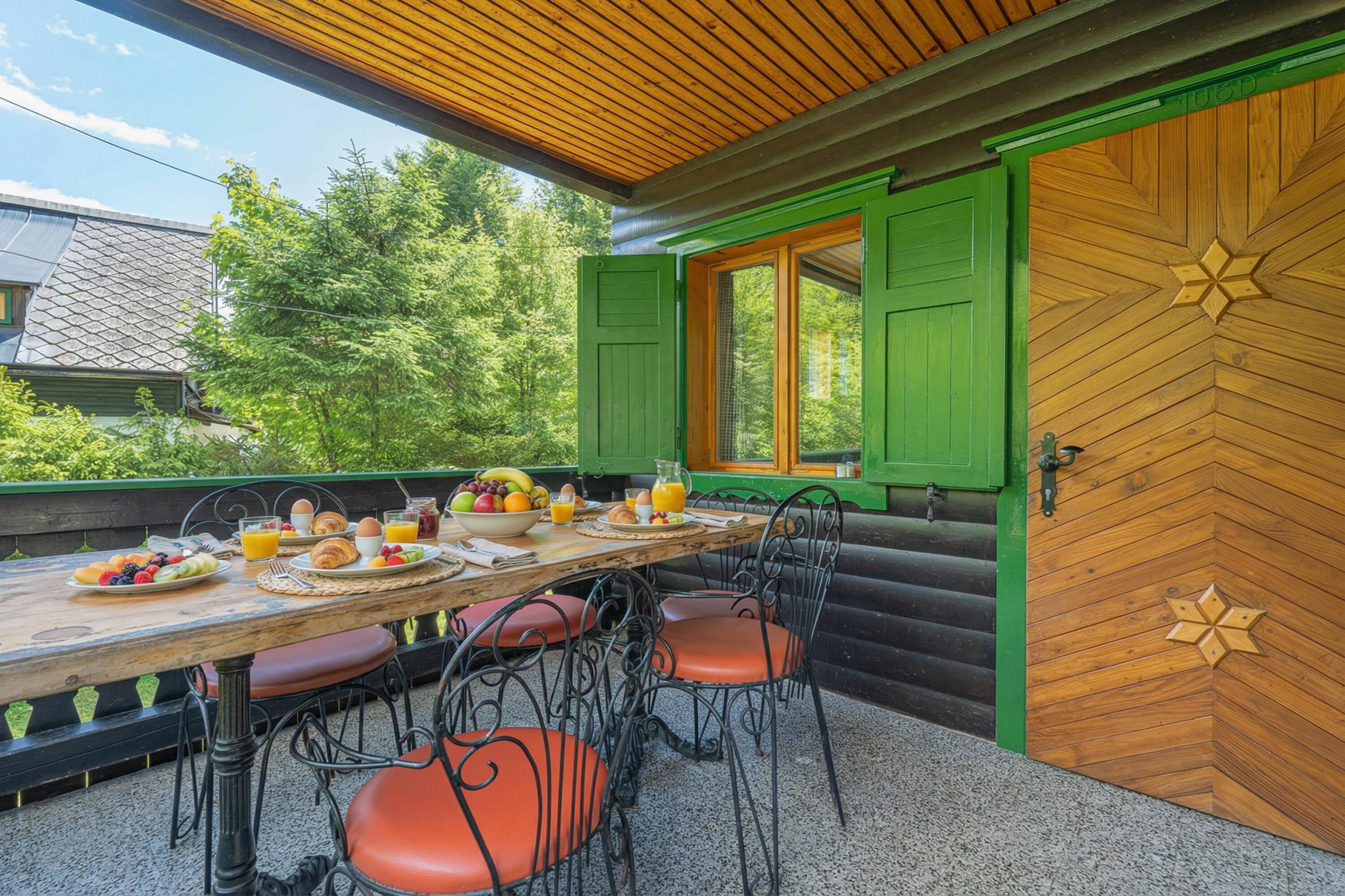 Una tavola di legno apparecchiata per la colazione, circondata da sedie, con vista su piante e una casa sullo sfondo.