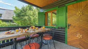 Una tavola di legno apparecchiata per la colazione, circondata da sedie, con vista su piante e una casa sullo sfondo.