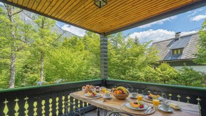A wooden table set with breakfast items, surrounded by chairs, on a balcony with a view of trees and a house. - Alpine Chalet Metka at Lake Bohinj (Bohinj)