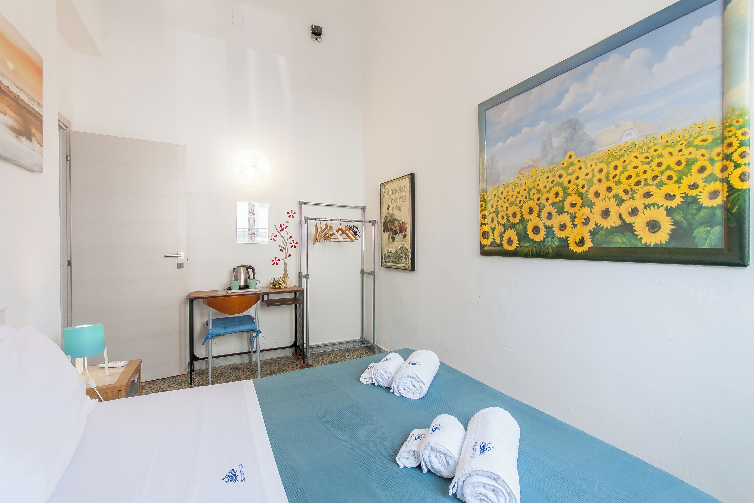 A hotel room with a bed, a desk with a chair, a sink, and a painting of sunflowers.
