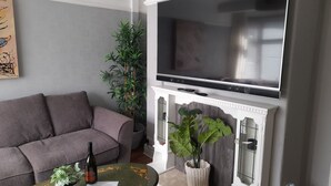 Un salon avec un canapé gris, une table ronde recouverte d’une nappe à motifs floraux, un téléviseur fixé au-dessus d’une cheminée et deux plantes en pot.