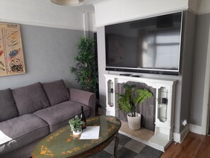 A living room with a grey sofa, a glass coffee table with a floral pattern, a flat-screen TV mounted above a white fireplace, and a large green plant beside the fireplace.