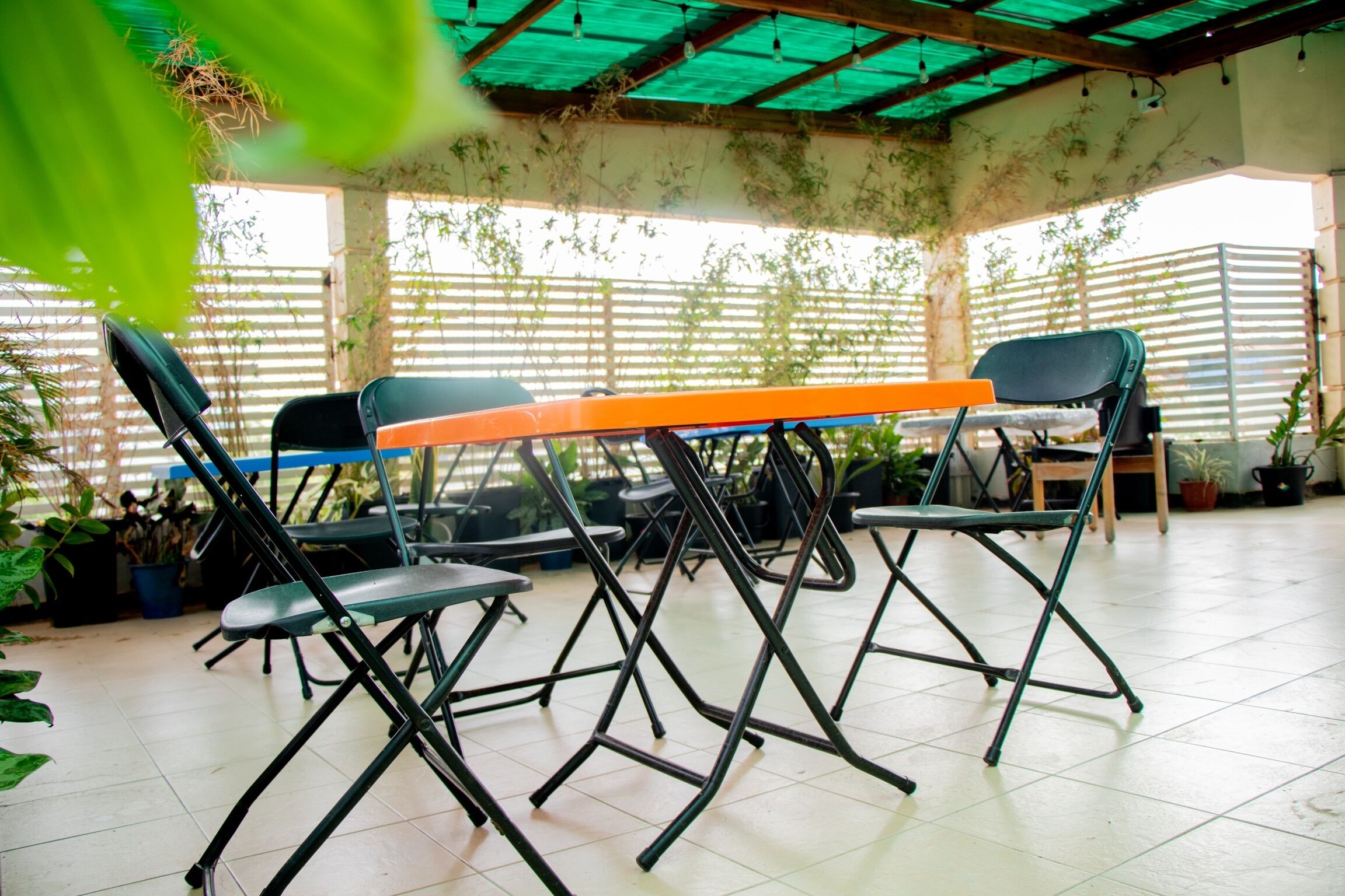 Een overdekt terras met opklapbare stoelen en een tafel, omgeven door potplanten en een bamboomscherm.