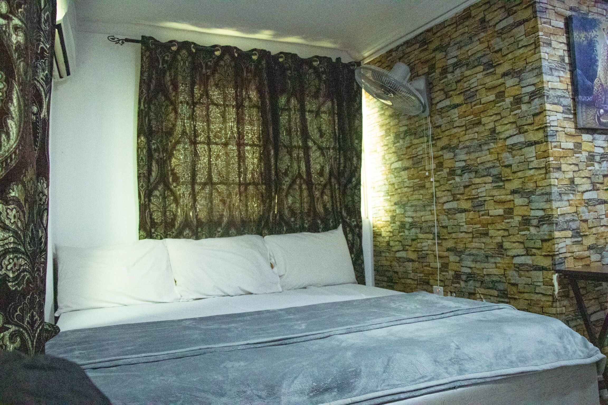 A bed with white linens, a dark patterned curtain, a wall fan, and a stone wall.
