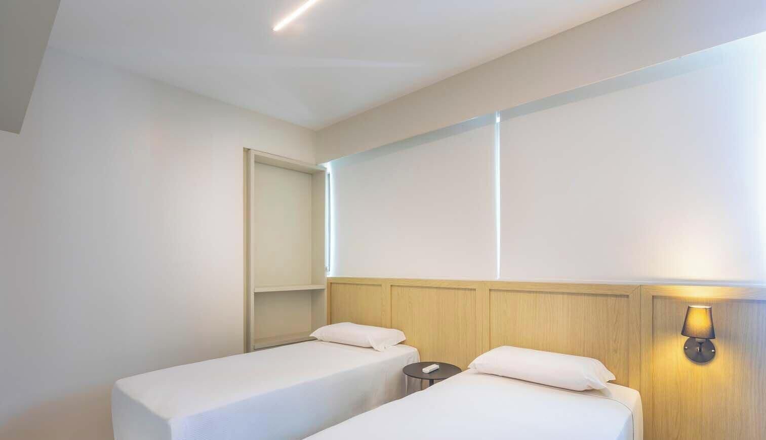 Une chambre d’hôtel avec deux lits simples, une table de chevet, une lampe fixée au mur et une porte.
