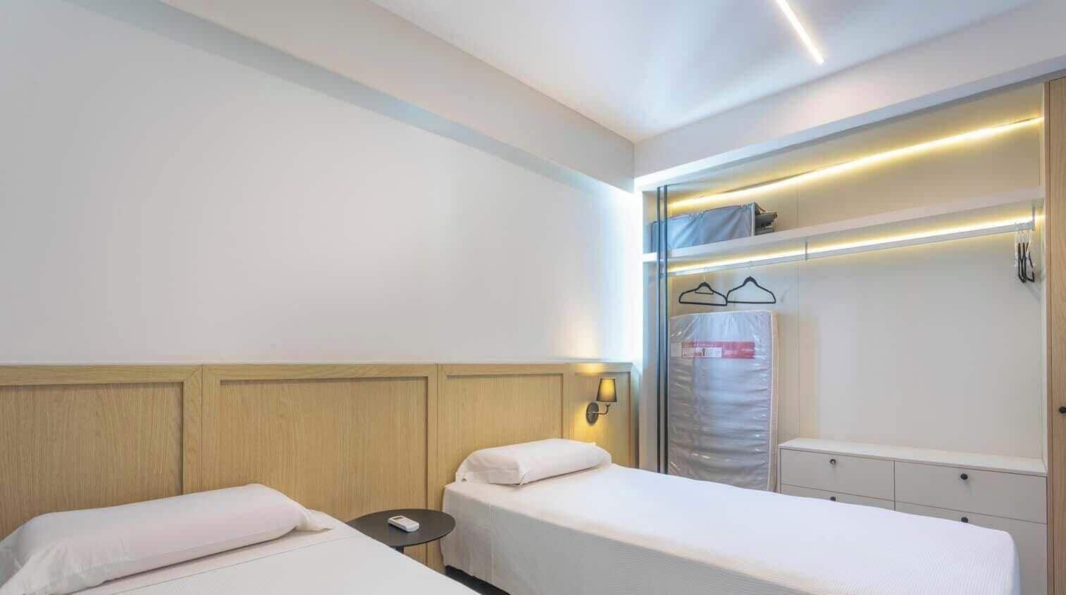 A hotel room with two single beds, a bedside table, a small round table, and a wardrobe with a mirror.