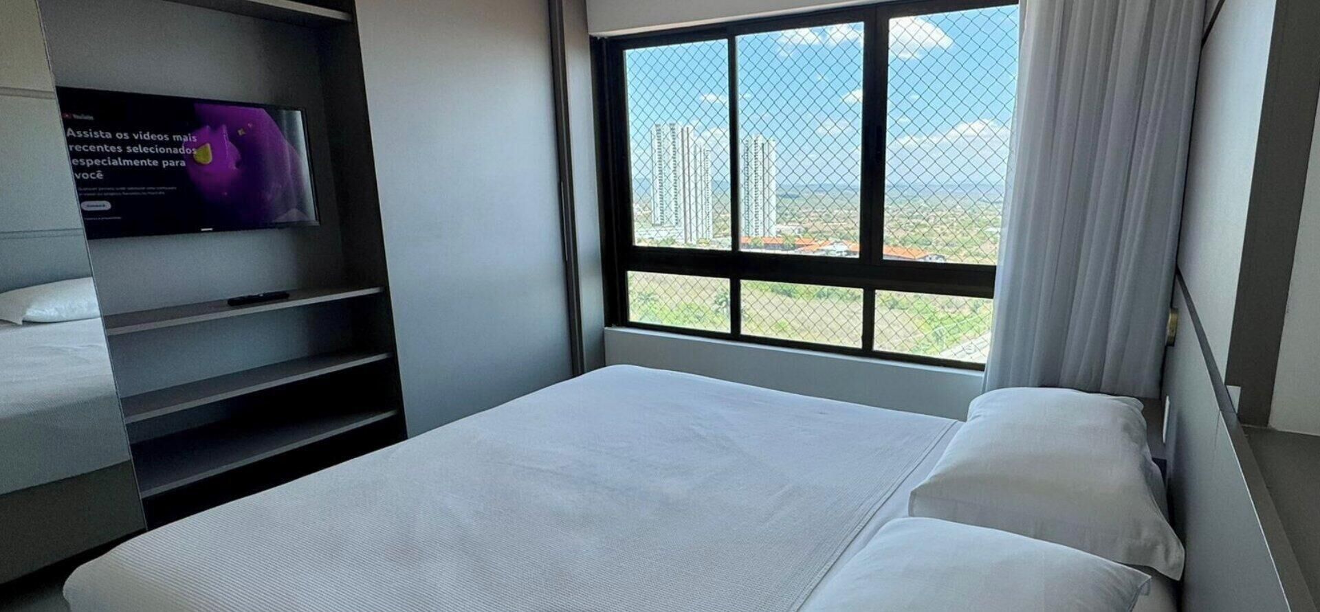 A modern hotel room with a bed, a TV on a stand, and a large window offering a city view.
