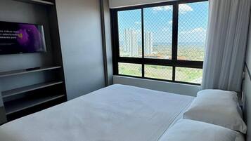 A modern hotel room with a bed, a TV on a stand, and a large window offering a city view.