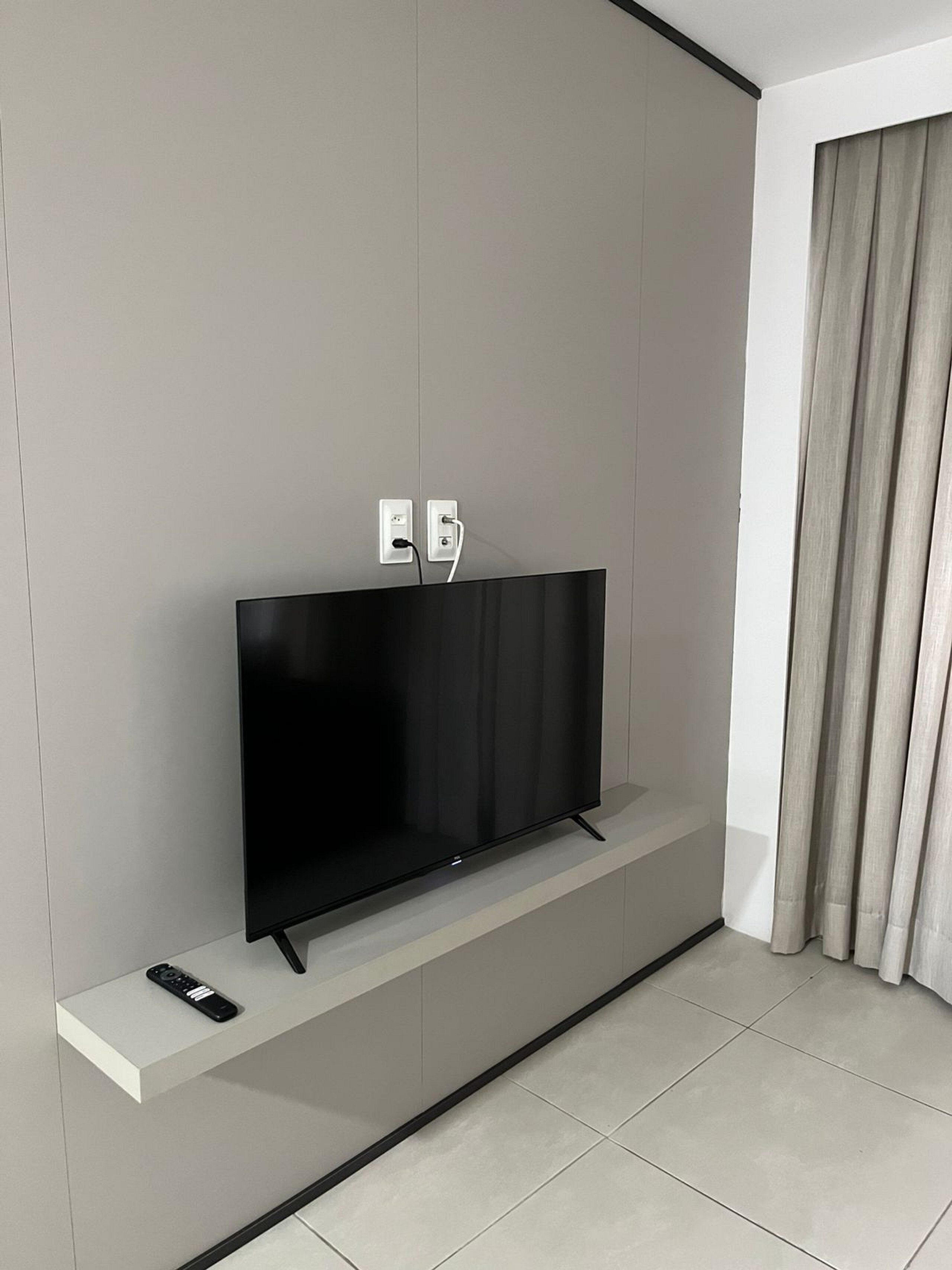 En moderne stue med et fladskærms-tv monteret på en hvid væg, en fjernbetjening på en hylde nedenunder, og et vindue med gardiner.