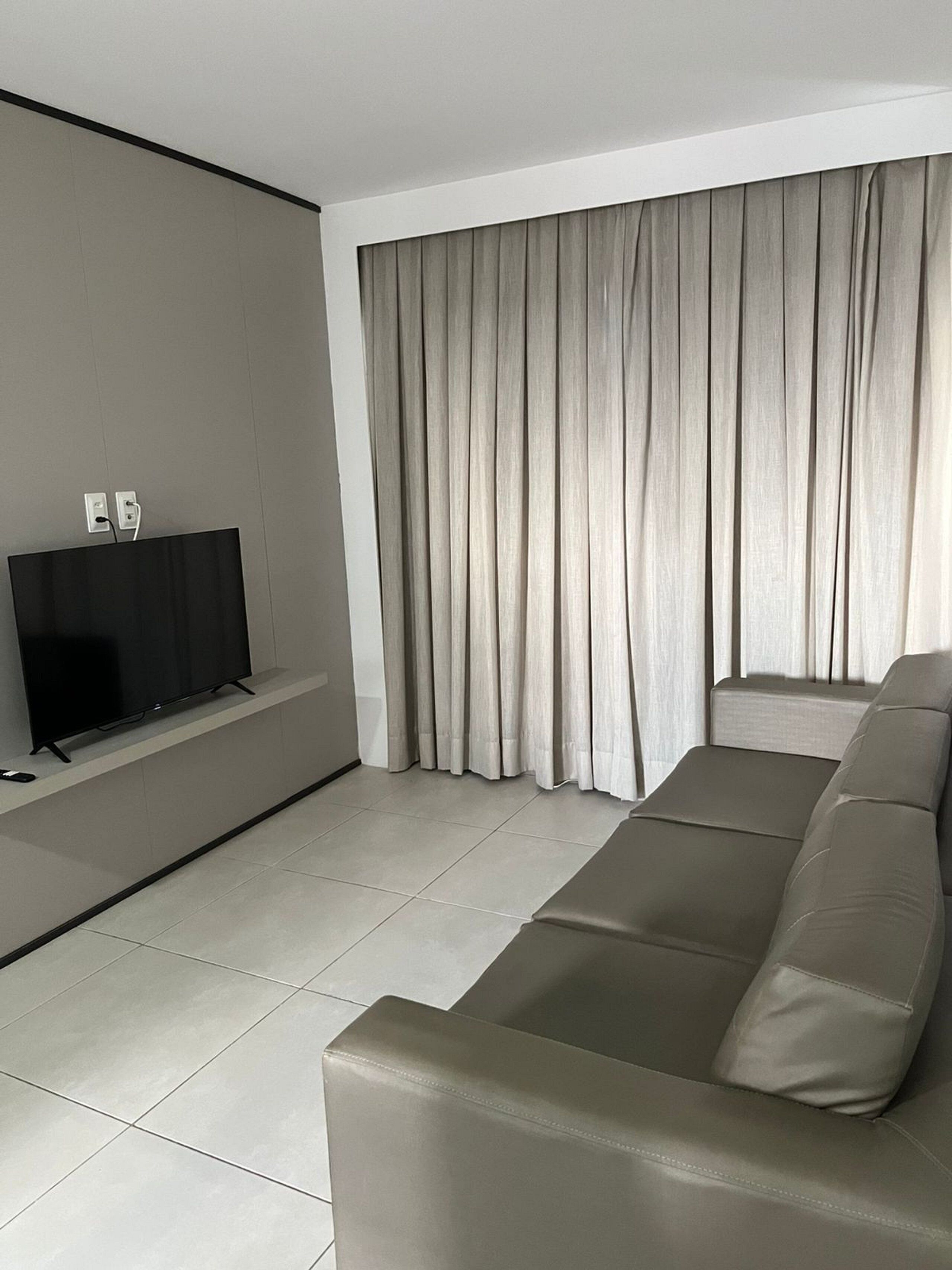 Una sala de estar moderna con una televisión de pantalla plana sobre un mueble bajo, un sofá grande de color gris y una ventana con cortinas grises.