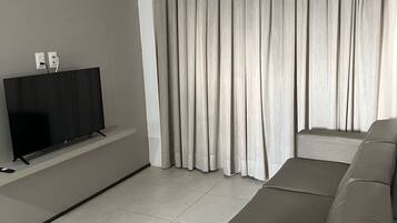 Una sala de estar moderna con una televisión de pantalla plana sobre un mueble bajo, un sofá grande de color gris y una ventana con cortinas grises.