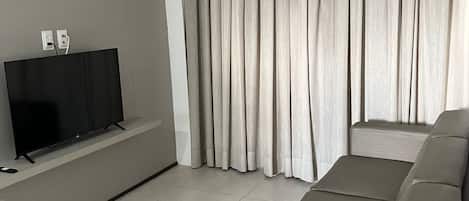 Ein modernes Wohnzimmer mit einem Flachbildfernseher auf einem Schrank, einem großen grauen Sofa und einem Fenster mit grauen Vorhängen.