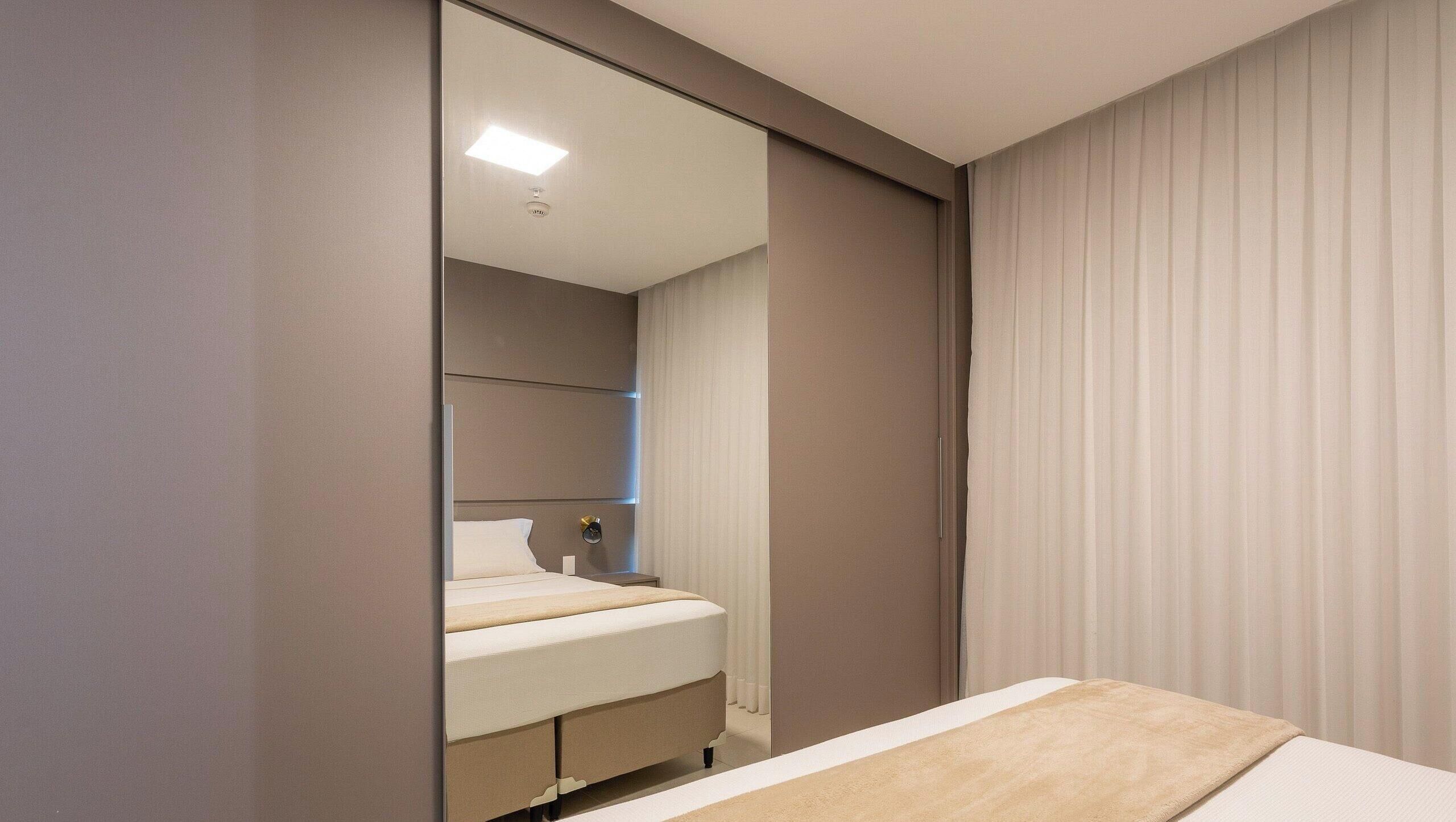 Un dormitorio con cama, mesita de noche, espejo y ventana con cortinas.