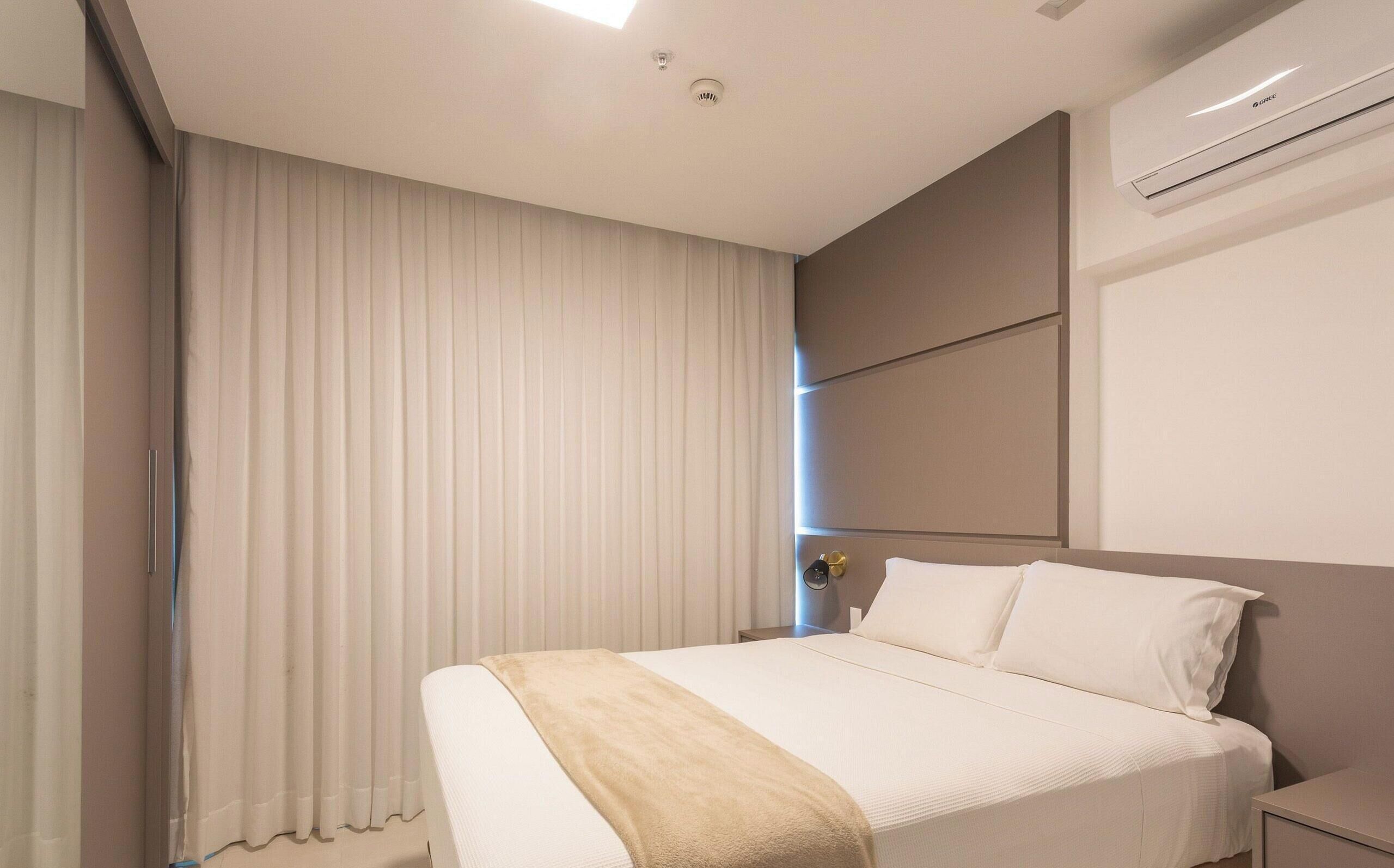 Una camera d'albergo con un letto, un comodino, un condizionatore d'aria a muro e una finestra con tende.