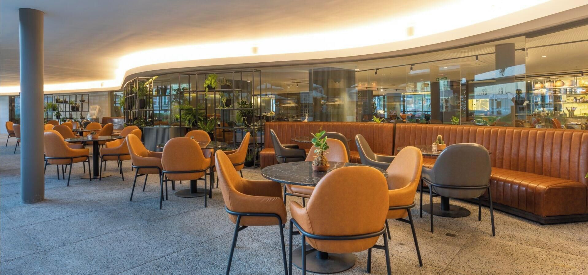 Un área de estar interior moderna con mesas y sillas redondas, una pared curva con una partición de vidrio y una cabina de cuero.