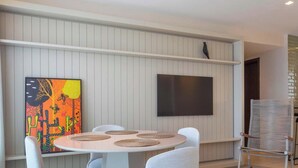 A modern dining area with a round table, white chairs, a flat-screen TV mounted on the wall, and a colorful abstract painting on the left. - Complexo Heron Marinho Flats (Campina Grande)