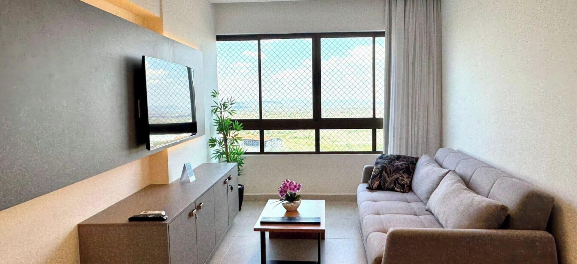 Ein stilvolles Wohnzimmer mit einer Couch, einem Couchtisch, einem an der Wand befestigten Fernseher und einem großen Fenster mit Ausblick.