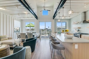 A modern kitchen with a large island, bar stools, and a dining area with chairs and a table. - Unique Coastal Container Home with Pool (Port Aransas)