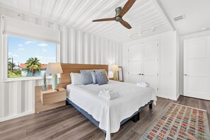 A bedroom with a bed, bedside lamps, a ceiling fan, and a view of the outdoors. - Unique Coastal Container Home with Pool (Port Aransas)
