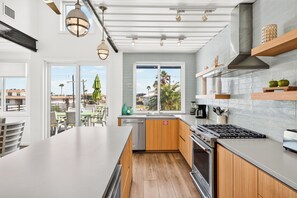 A modern kitchen with a large island, wooden cabinets, and a view of palm trees outside. - Unique Coastal Container Home with Pool (Port Aransas)