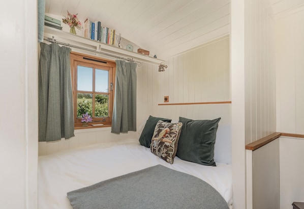 A bedroom with a bed, a window with curtains, a shelf with books and a clock, and a view of greenery outside.