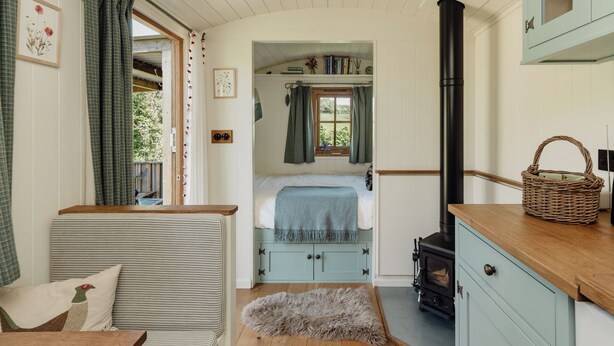 A cozy room with a bed, a fireplace, a window with curtains, a wooden bench, and a basket on the counter. - Hurdlemakers Hut (Dorchester)