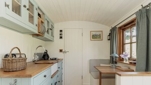 A compact kitchen with light blue cabinets, a wooden countertop, a bench, and a window with curtains. - Hurdlemakers Hut (Dorchester)