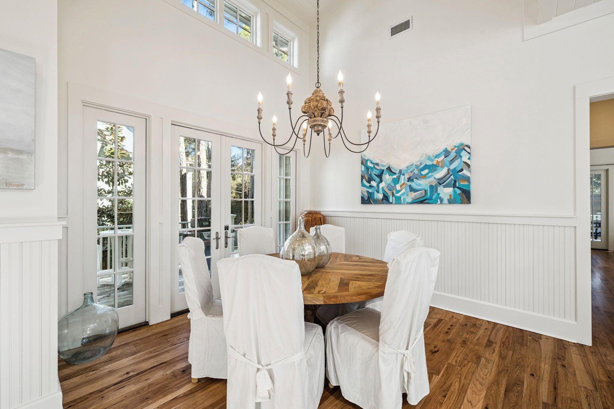 A dining area with a wooden table, white chairs, a chandelier, and a large abstract painting on the wall.