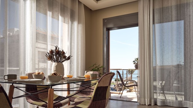 A dining area with a glass table, chairs, and a view of the sea through large windows. - Laura Suites (Zakynthos)