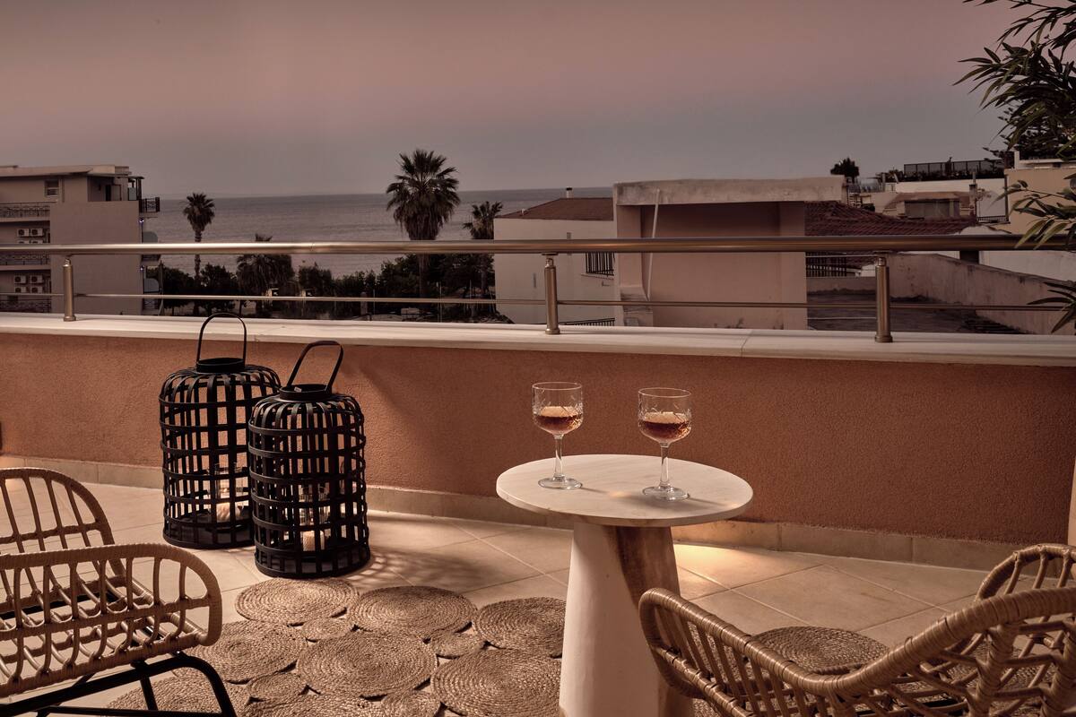 A terrace with wicker furniture, two wine glasses, and a view of the sea and buildings.