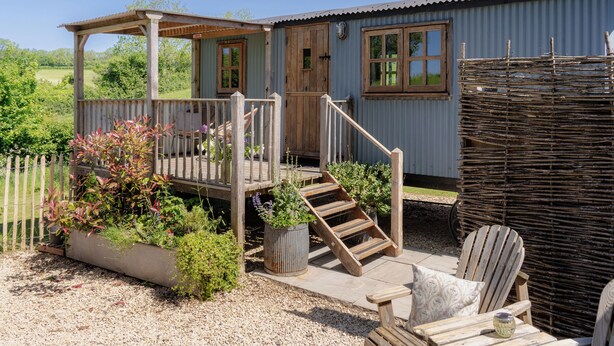 A rustic cabin with a wooden deck, a staircase, and a seating area with a chair and table. - Spindleberry Hut (Dorchester)