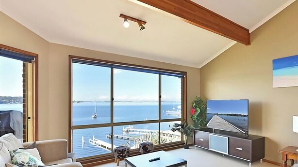 A living room with large windows, a flat-screen TV, a sofa, and a lamp. - Sparkling Waters - Jetty & Games Room! (Rathmines)