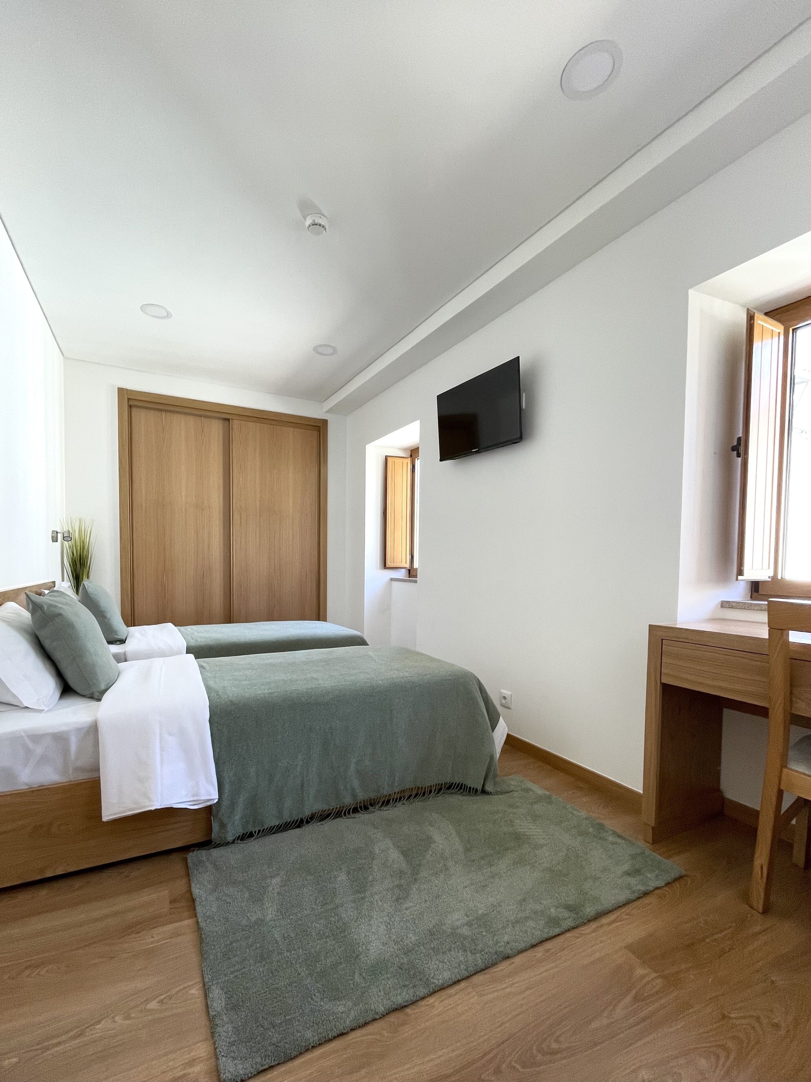 A modern bedroom with two beds, a desk, a TV, and a window.