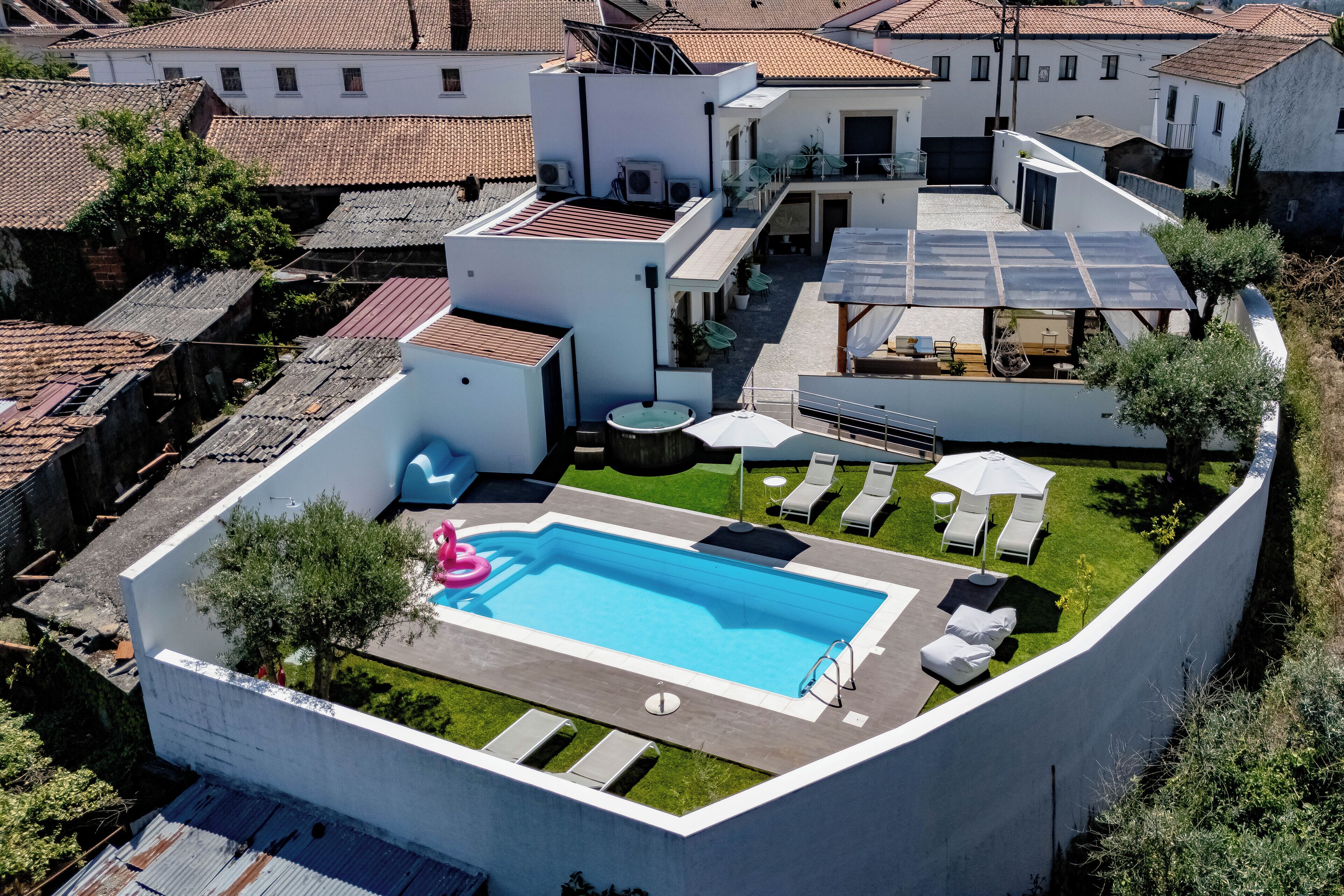 Una piscina en la azotea con sillones de descanso y sombrillas, rodeada por un muro blanco y follaje.