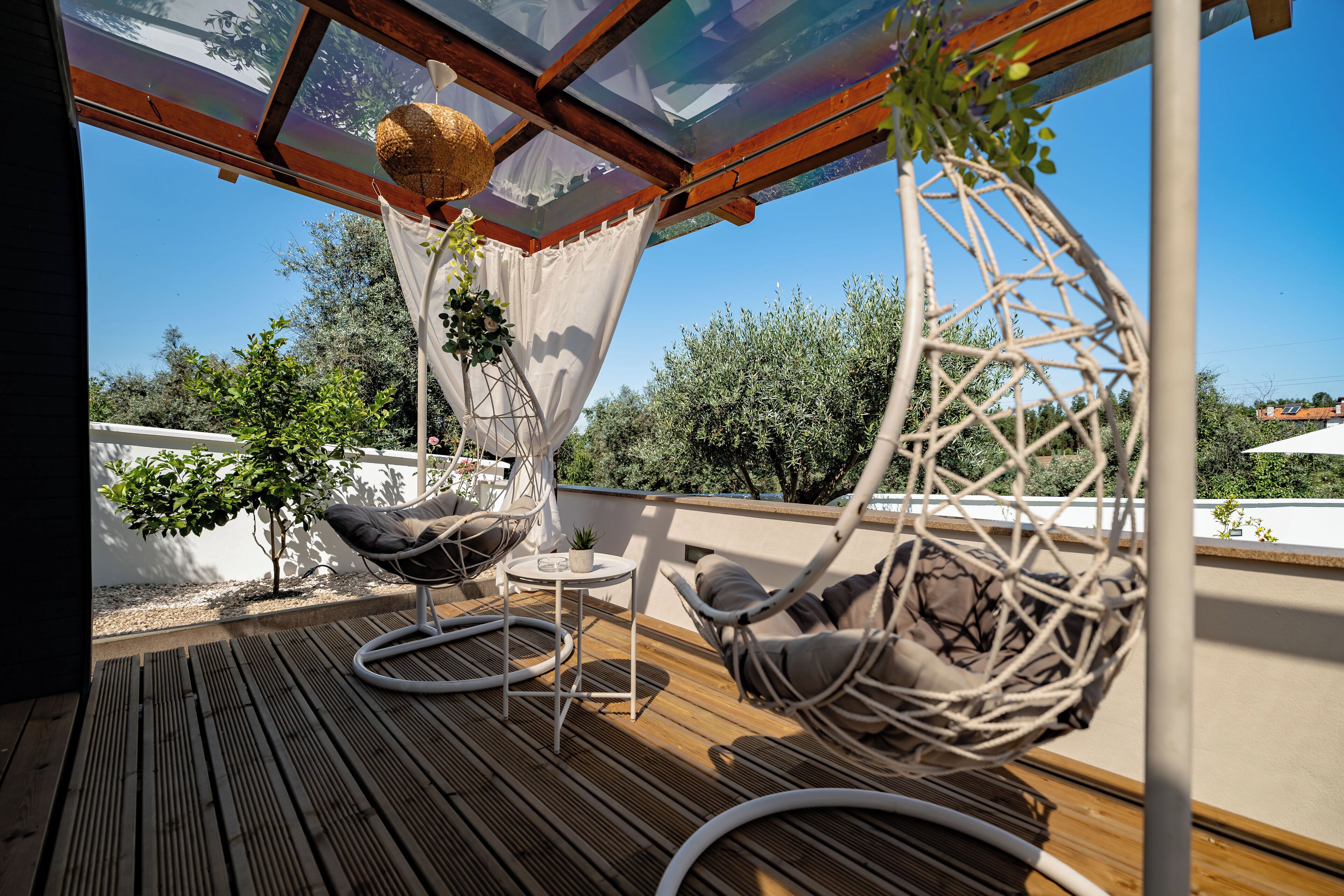 A wooden deck with two wicker chairs and a small table, overlooking a garden with trees and a clear sky.