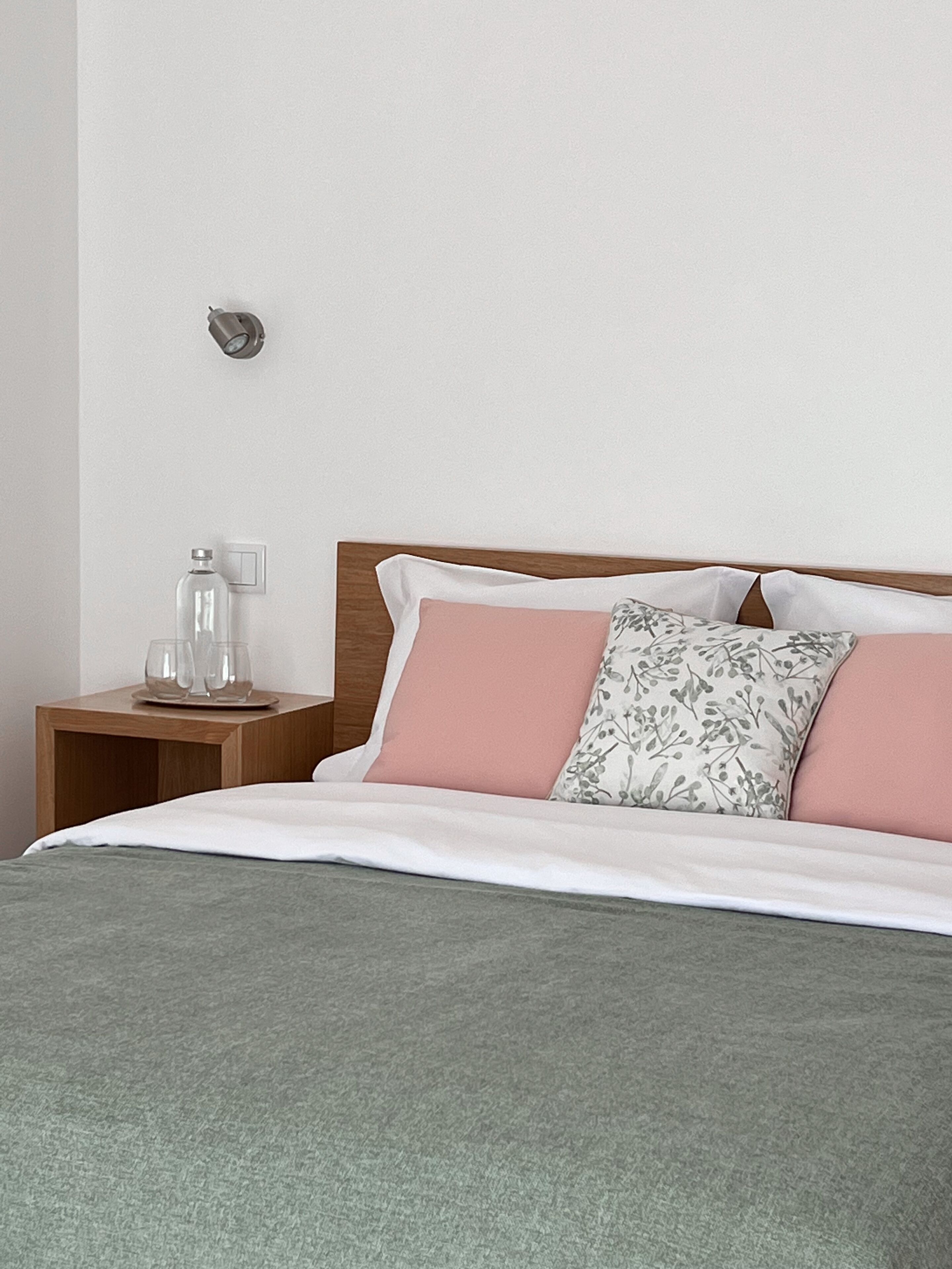 A neatly made bed with a grey blanket, a wooden nightstand with a glass bottle and two glasses, and a wall-mounted light fixture.