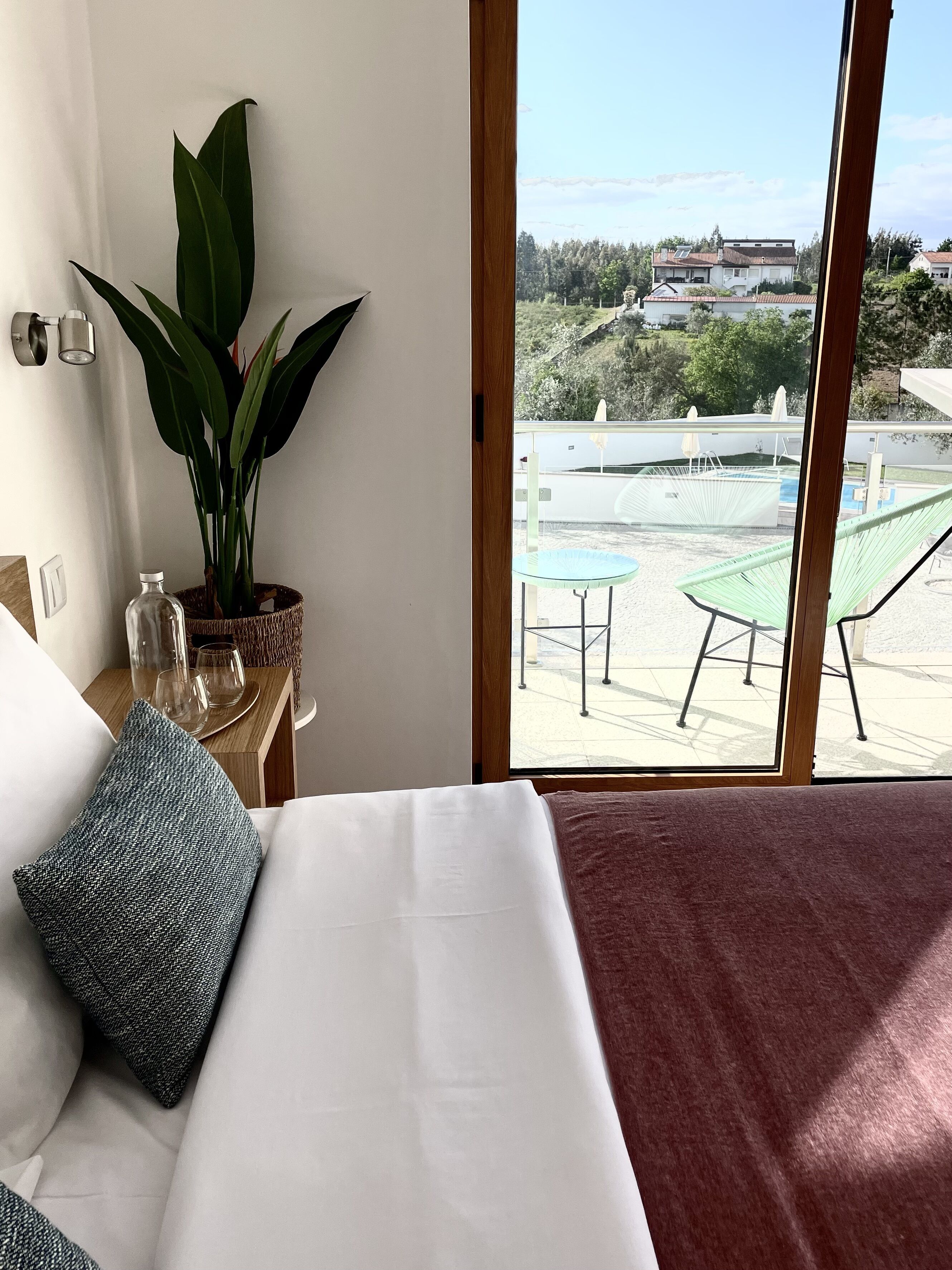 A bedroom with a large bed, a potted plant, and a view of a pool and houses through a window.