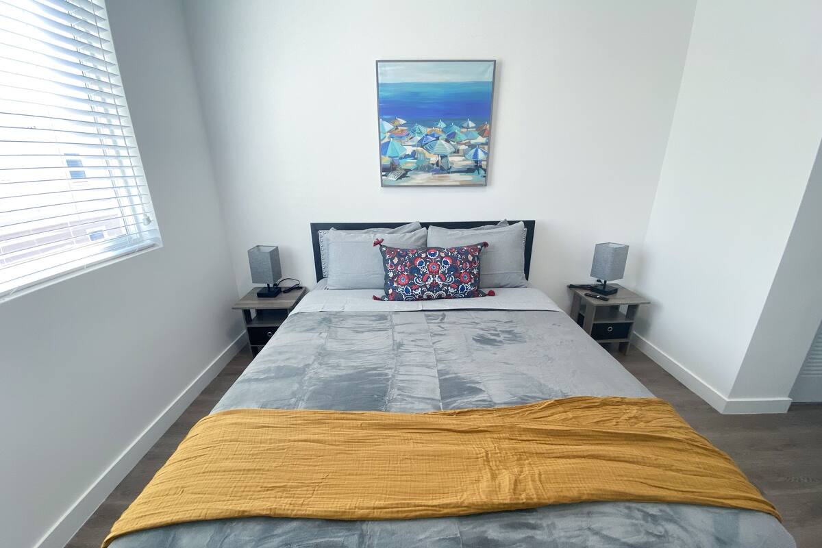 Un dormitorio con una cama grande, una mesita de noche con lámpara, un cuadro en la pared y una ventana con persianas.
