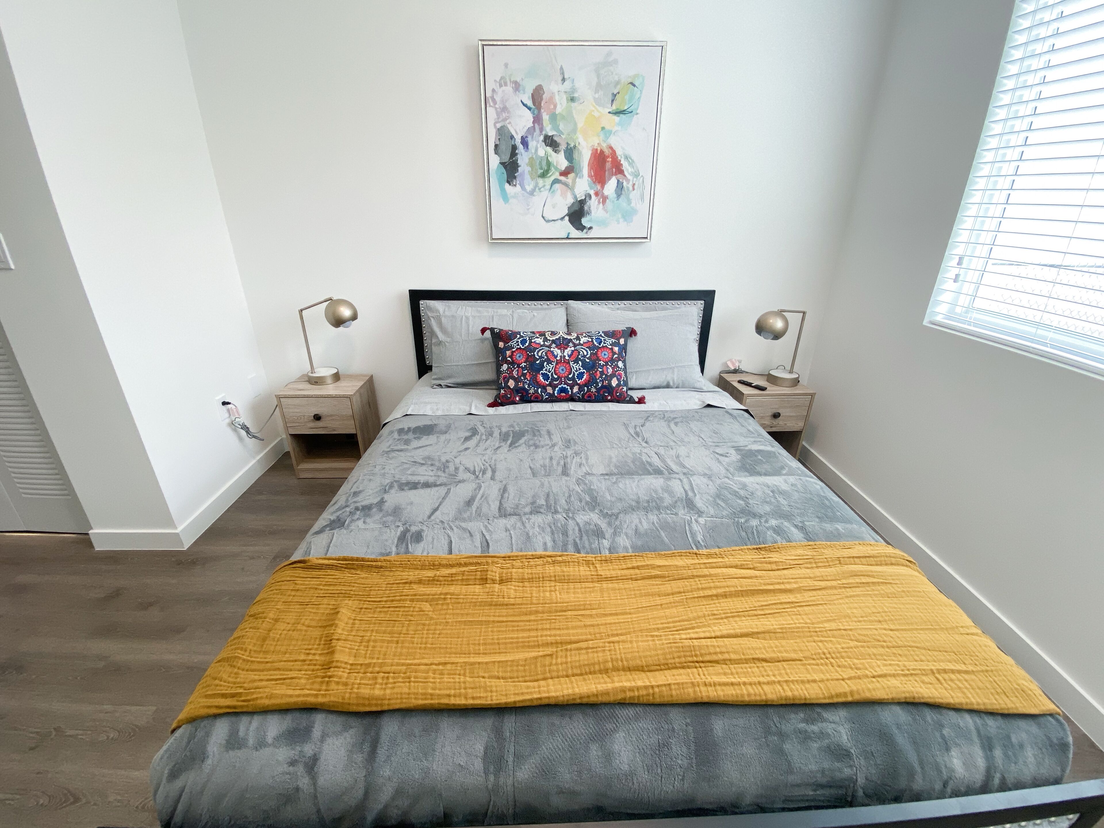 Ein Schlafzimmer mit einem Bett, zwei Nachttischen, einem Fenster mit Jalousien und einem an der Wand hängenden abstrakten Gemälde.