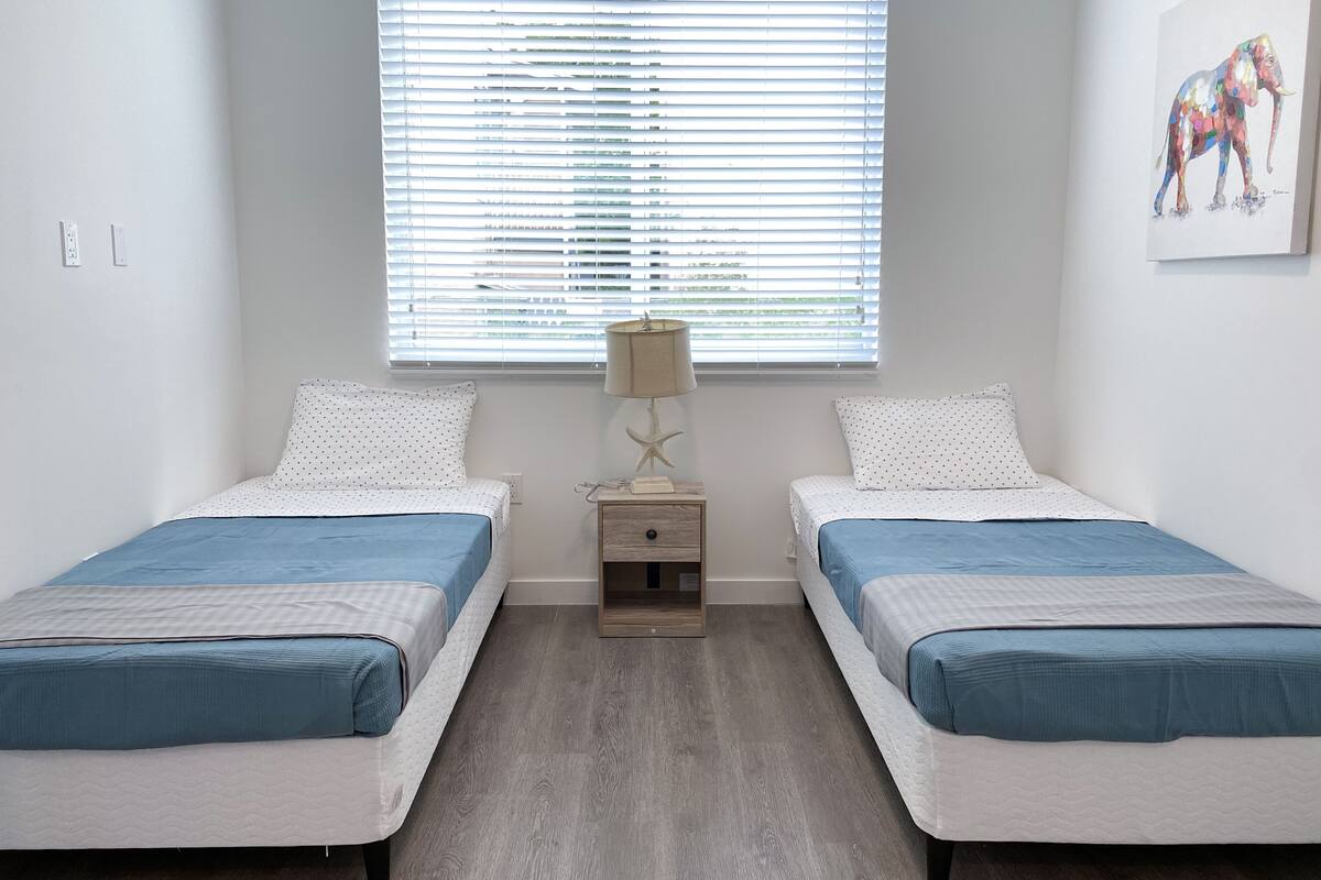 Un dormitorio con dos camas, una mesita de noche, una lámpara y una ventana con persianas.
