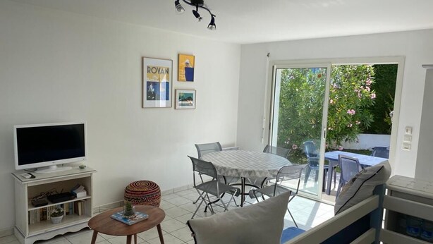 Uma sala de estar com televisão, mesa de jantar com cadeiras, sofá e varanda com vista para o verde.