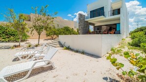 A modern two-story house with a balcony, white lounge chairs, and a sandy landscape. - Luxe Beachfront Villa, Private Pool & Beach Access (Progreso)