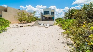 A modern beach house with a two-story structure, a sandy patio with lounge chairs, and a staircase leading to the upper level. - Luxe Beachfront Villa, Private Pool & Beach Access (Progreso)