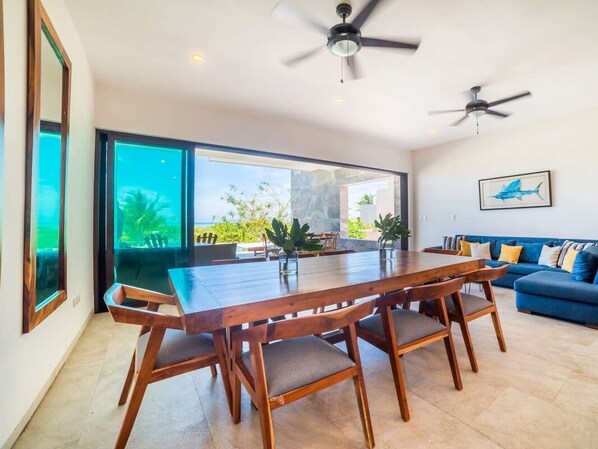 Una habitación amplia con un gran comedor, dos ventiladores de techo, un sofá azul y un ventanal que ofrece vistas a zonas verdes.