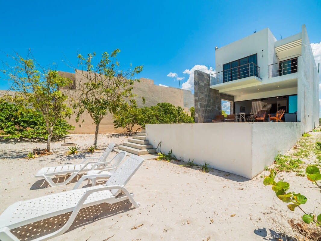 A modern two-story house with a balcony, white lounge chairs, and a sandy landscape.
